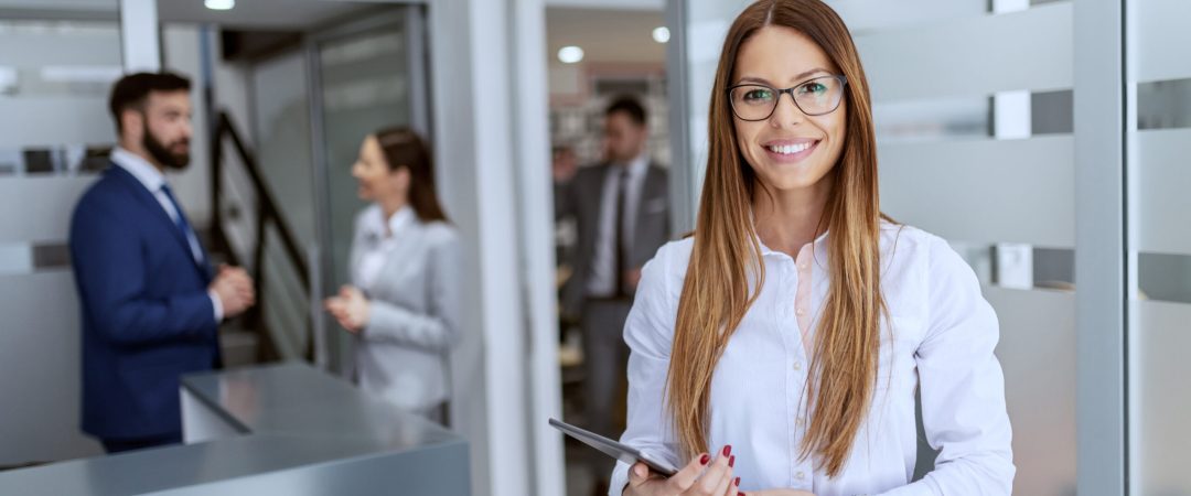 Charming Caucasian businesswoman dressed in shirt and skirt standing on hallway and holding tablet. In background her colleagues standing and chatting.